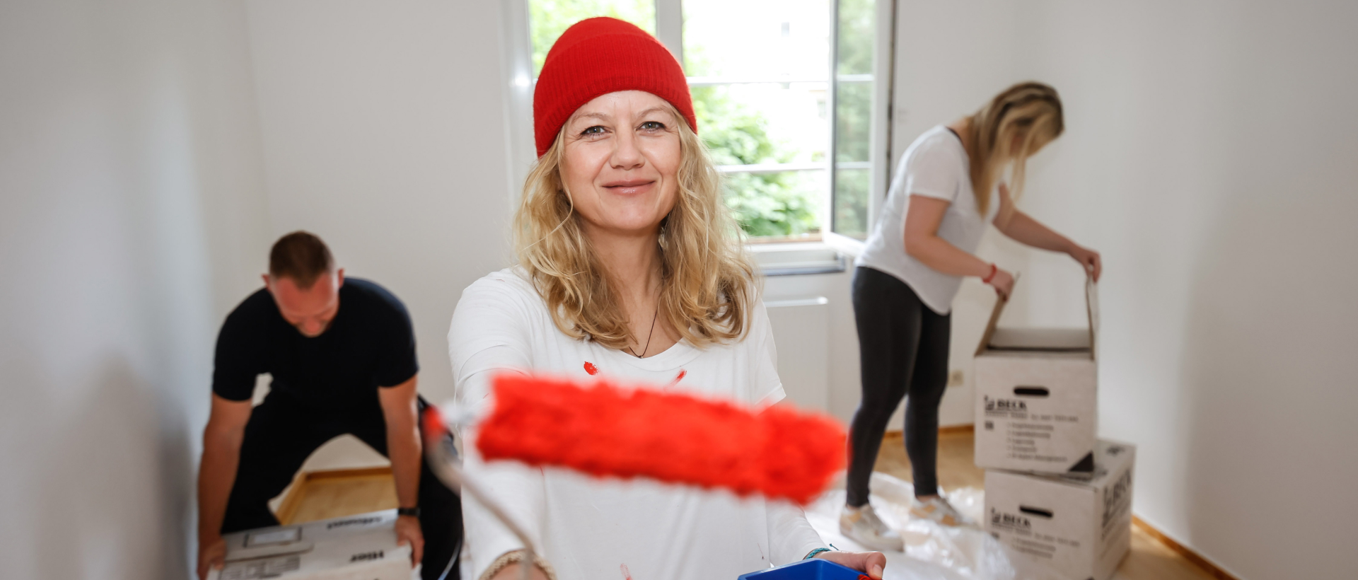 Lächelnde Frau mit roter Mütze hält eine Malerolle mit roter Farbe in die Kamera.