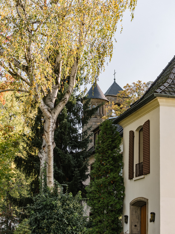 Auf dem Bild sieht man einen Baum neben einem Haus und im Hintergrund erkennt man 2 Kirchtürme 