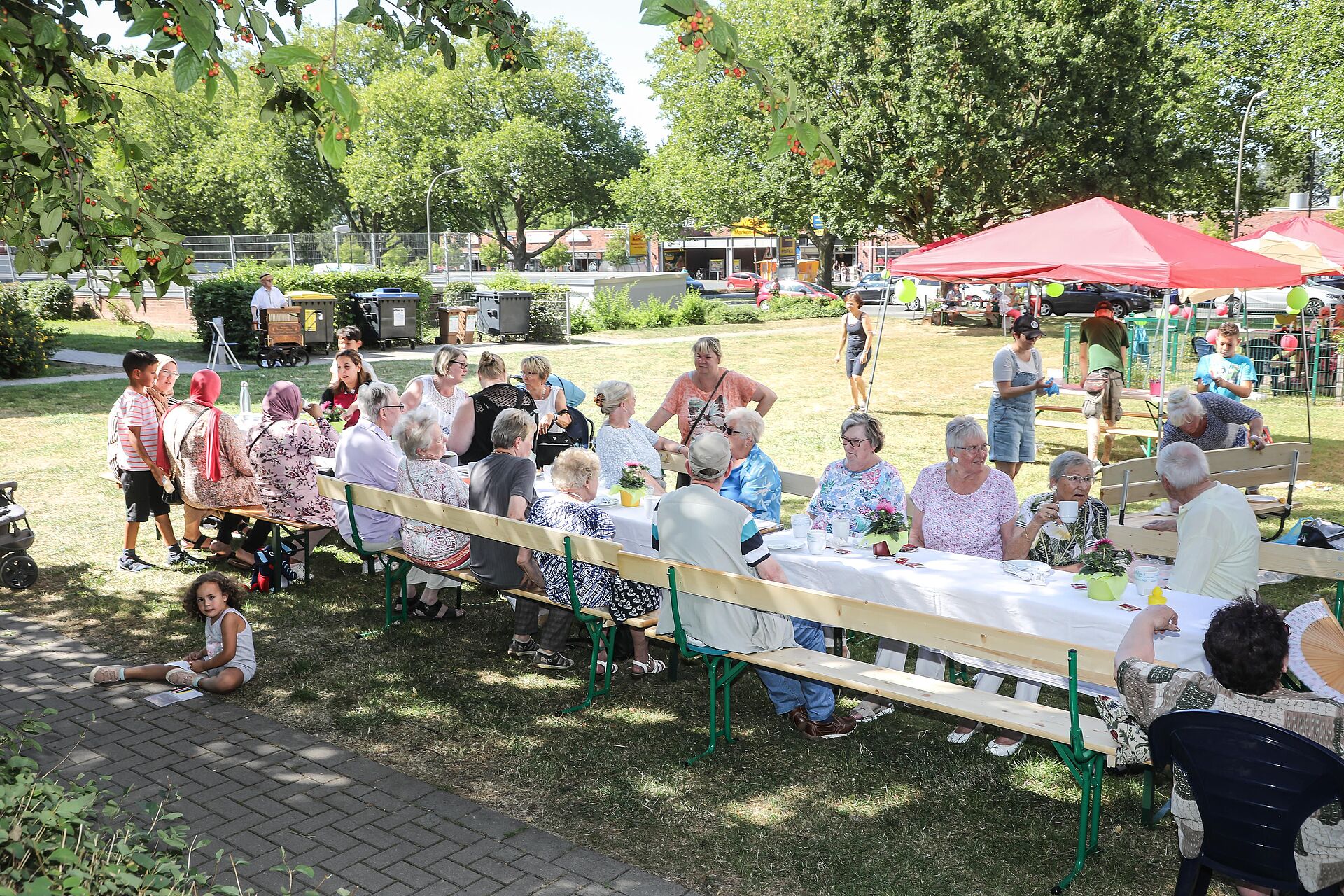 Nachbarinnen und Nachbarn sitzengemeinsam an einer langen Kaffeetafel im schattigen Garten der Nachbarschaftsagentur