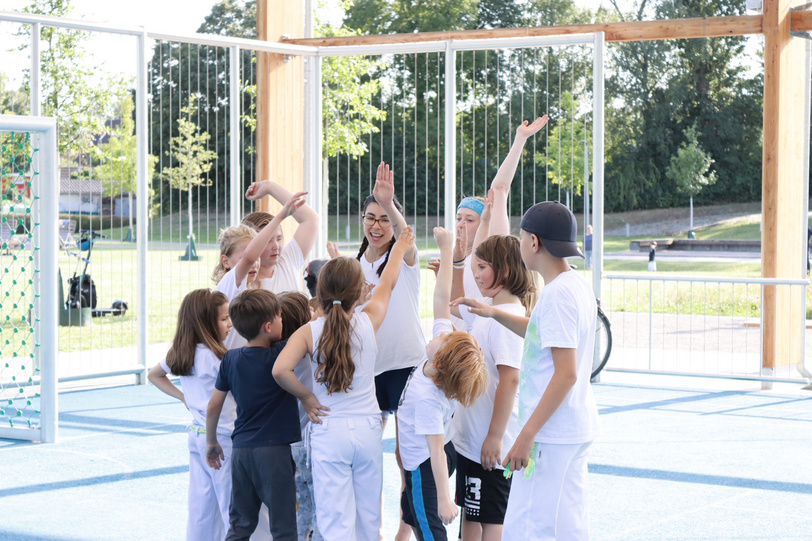 Kinder und Jugendliche in Sportbekleidung stehen im Kreis auf einer Spielfläche in Park und klatschen sich zur Begrüßung ab.