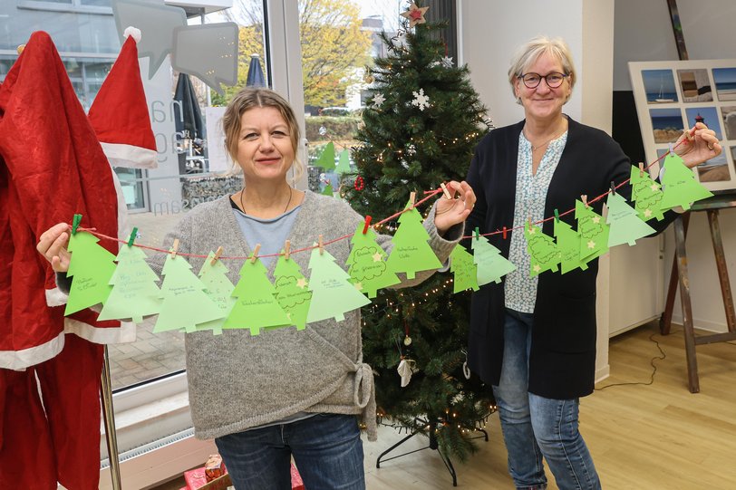 Zwei Frauen stehen in einem hellen, gemütlichen Raum und halten Papiergirlanden mit Geschenkewünschen in Richtung des Betrachters