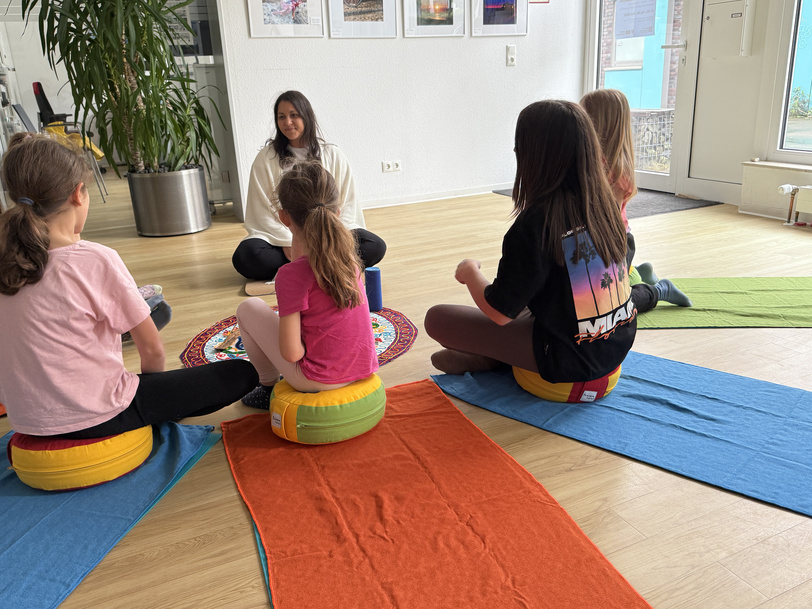 Mehrere Menschen sitzen auf Kissen und Yogamatten in einem hellen Raum.