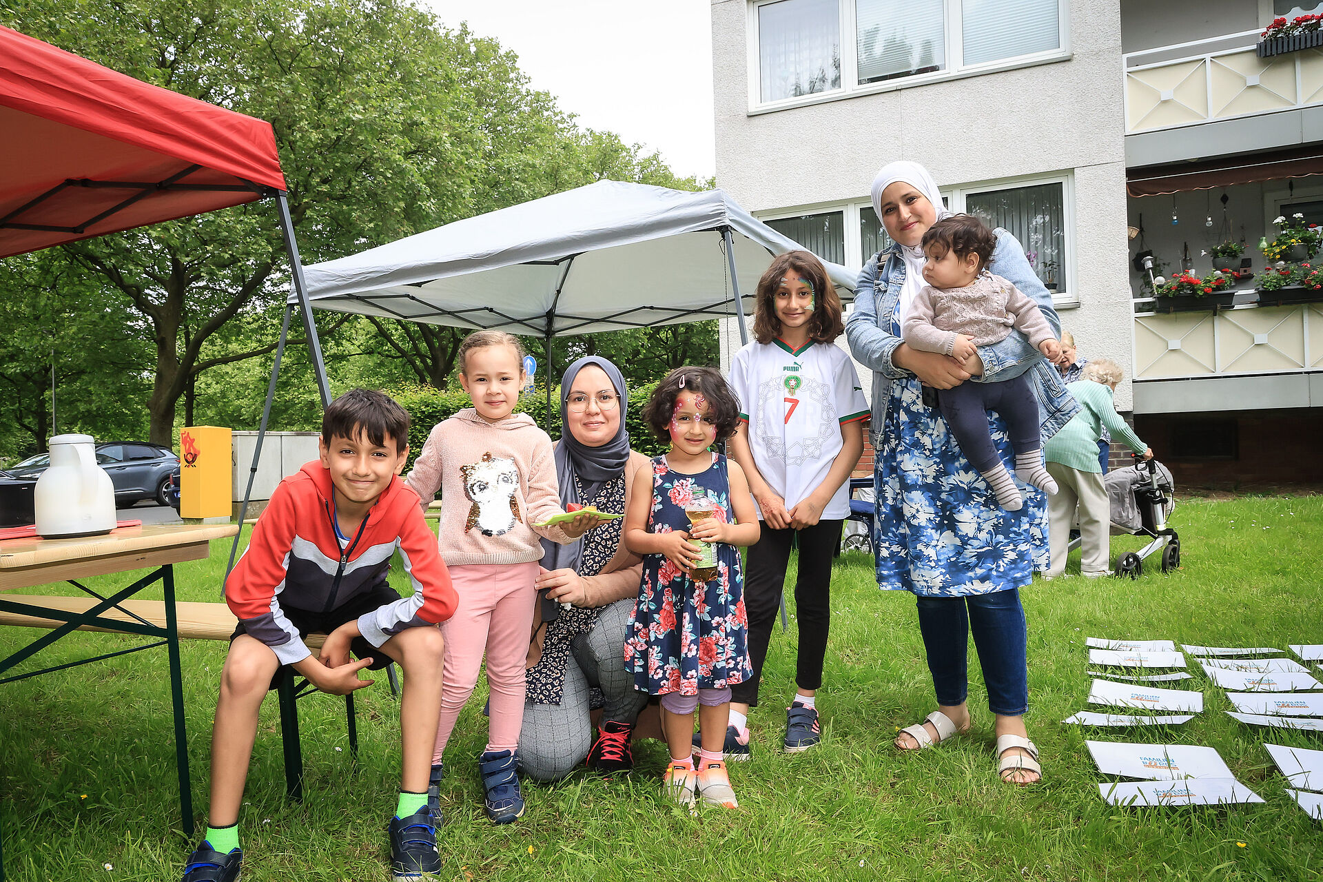 Zwei Frauen und mehrere Kinder lächeln gutgelaunt im Garten der Nachbarschaftsagentur in die Kamera des Fotografen.