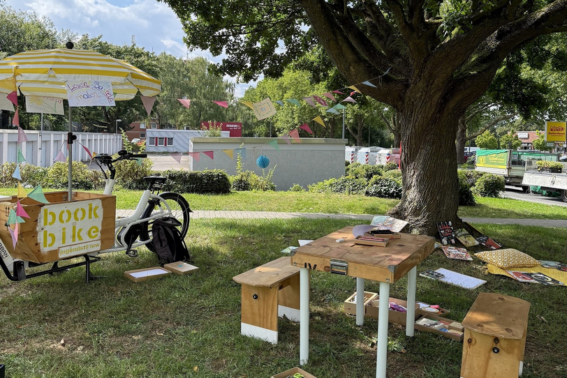 Auf der Wiese unter einem Baum steht ein Lastenfahrrad mit gelb-weißem Sonnenschirm, daneben gibt es Tisch und Bänke, eine Decke mit Kissen, Bücher und Bastelsachen.