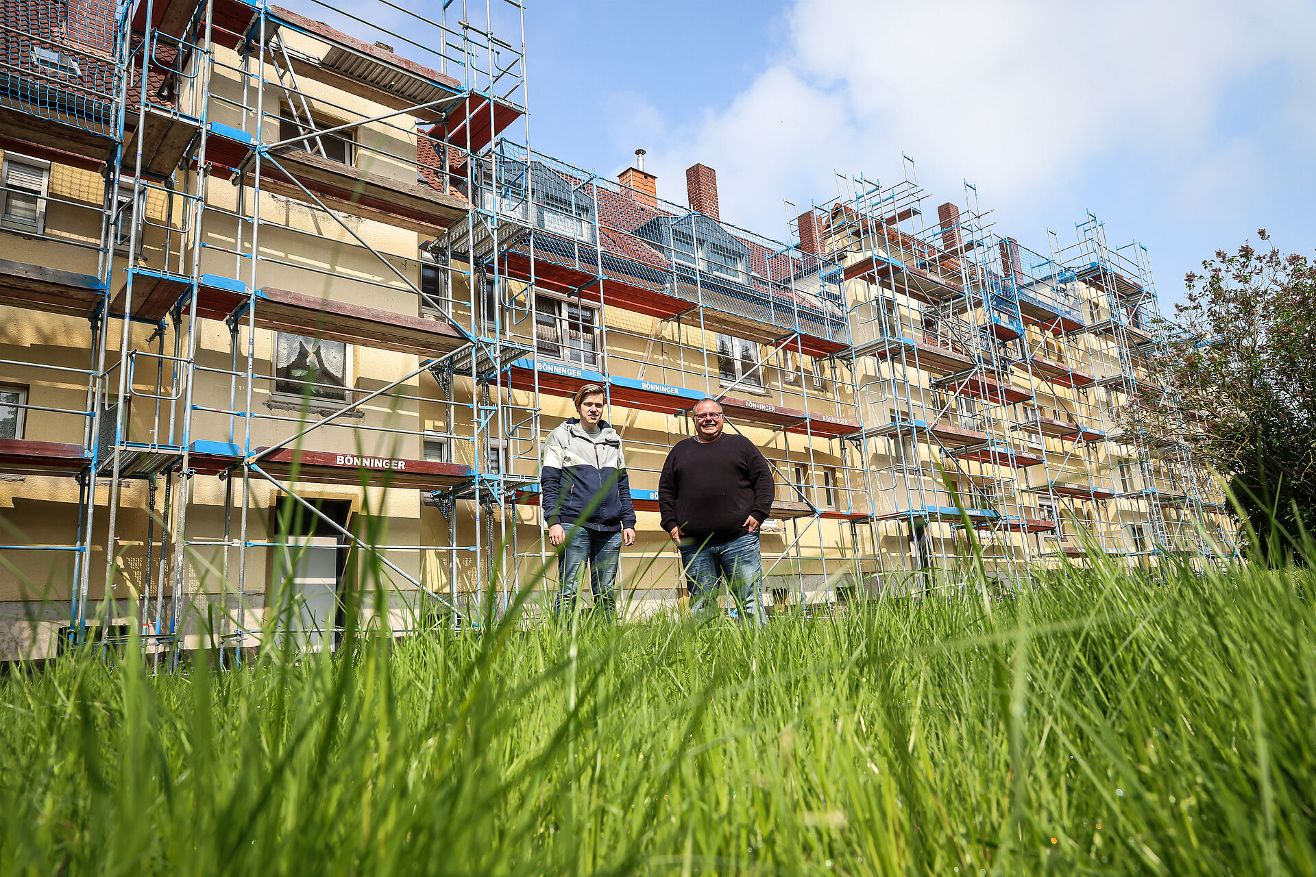 Zwei Männer, Bauleiter des Wohnungsunternehmens DOGEWO21, schauen in die Kamera, im Hintergrund stehen eingerüstete Häuser, die modernisiert werden