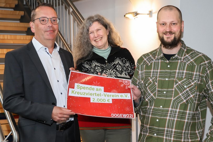 Zwei Männer und eine Frau stehen auf einer Holztreppe und zeigen einen symbolischen Spendenscheck im Wert von 2.000 Euro.