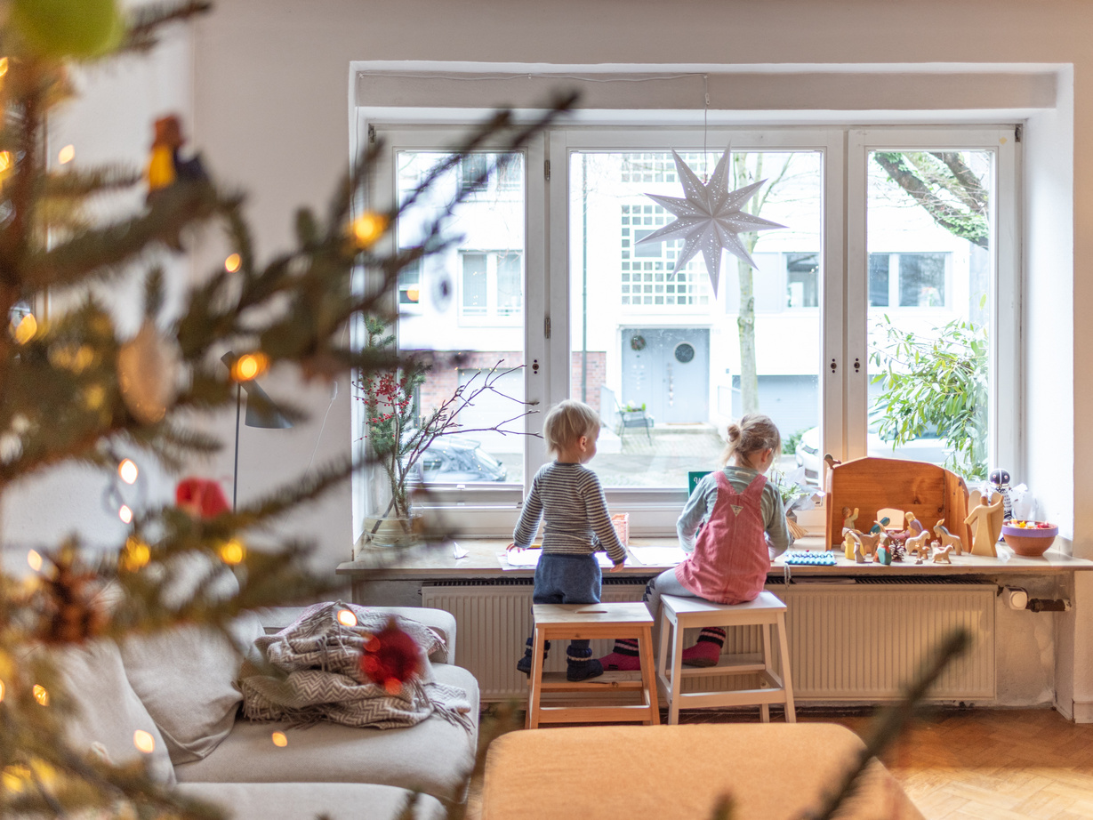 Zwei Kinder sitzen im weihnachtlich geschmückten Wohnzimmer auf einem Stuhl und schauen aus einem großen Fenster. 