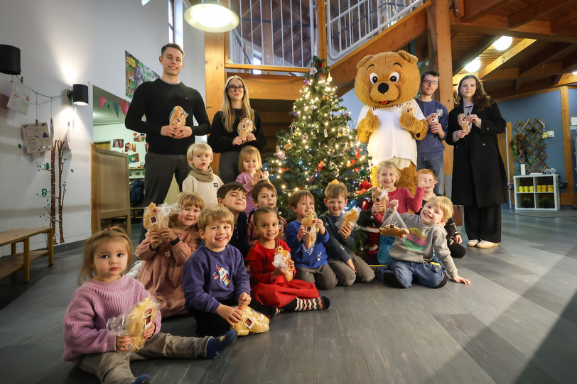 Vier junge Menschen, das Maskottchen Dogibär und eine Gruppe Kitakinder stehen und sitzen vor einem Weihnachtsbaum und halten Stutenkerle in den Händen.