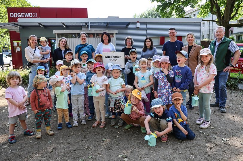 Viele kleine und große Menschen stehen auf einer Brachfläche vor einem DOGEWO21-Servicebüro mit Blumensamen und Kindergießkannen und lächeln in die Kamera.