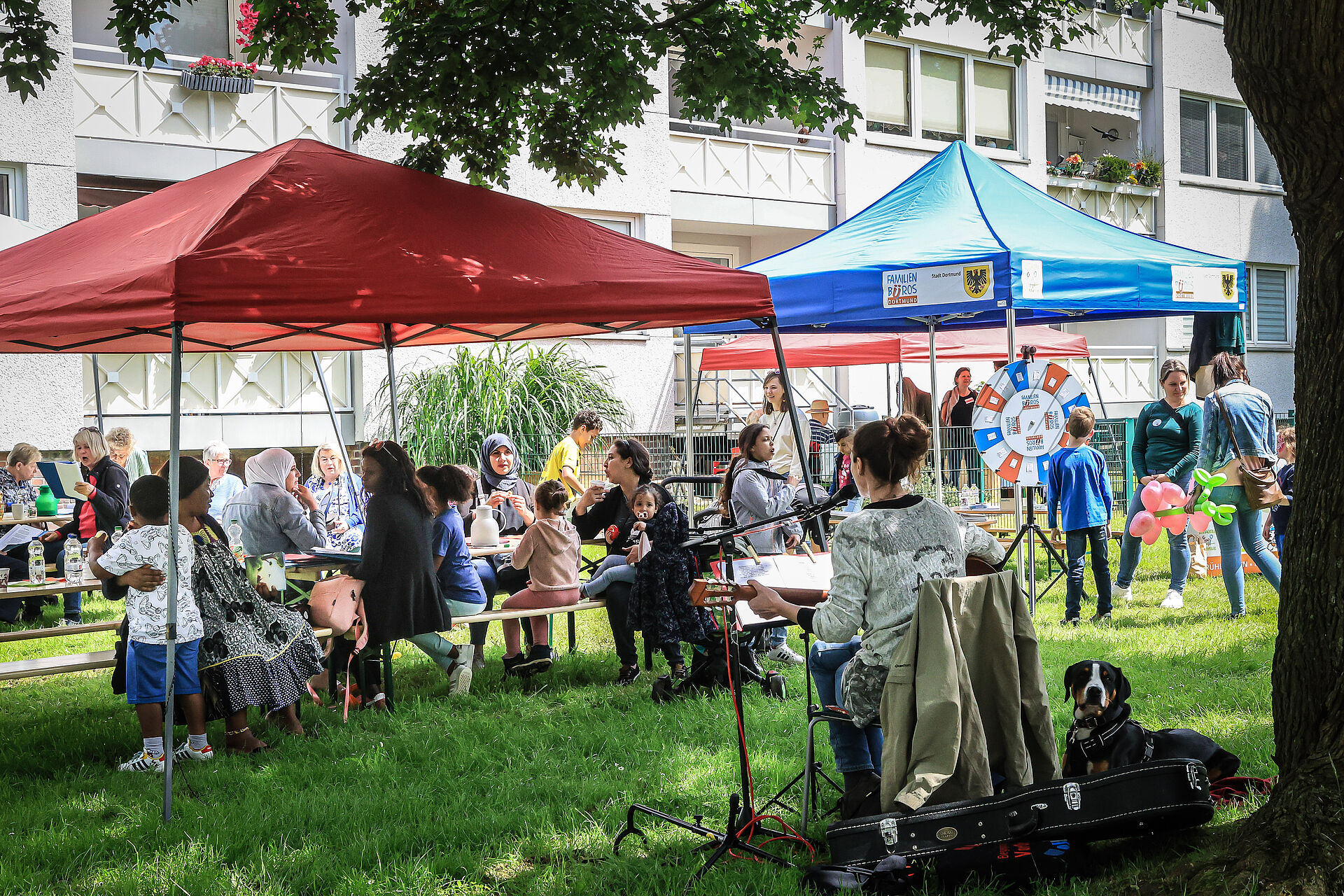 Menschen jeglichen Alters sitzen unter einem Pavillon und singen, essen und reden miteinander während eine Frau mit Gitaree im Vordergrund Lieder spielt.