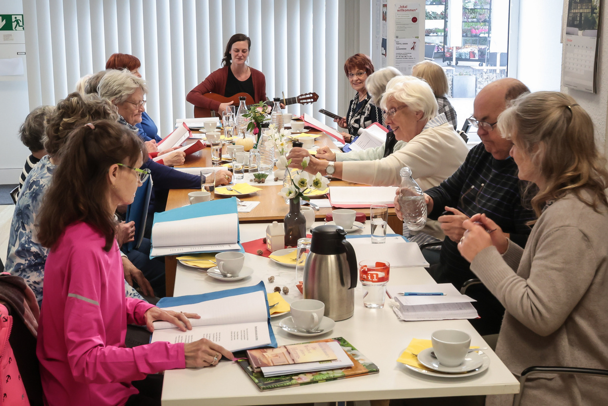Ein Dutzend Menschen sitzt um einen gedeckten Tisch und singt Lieder.