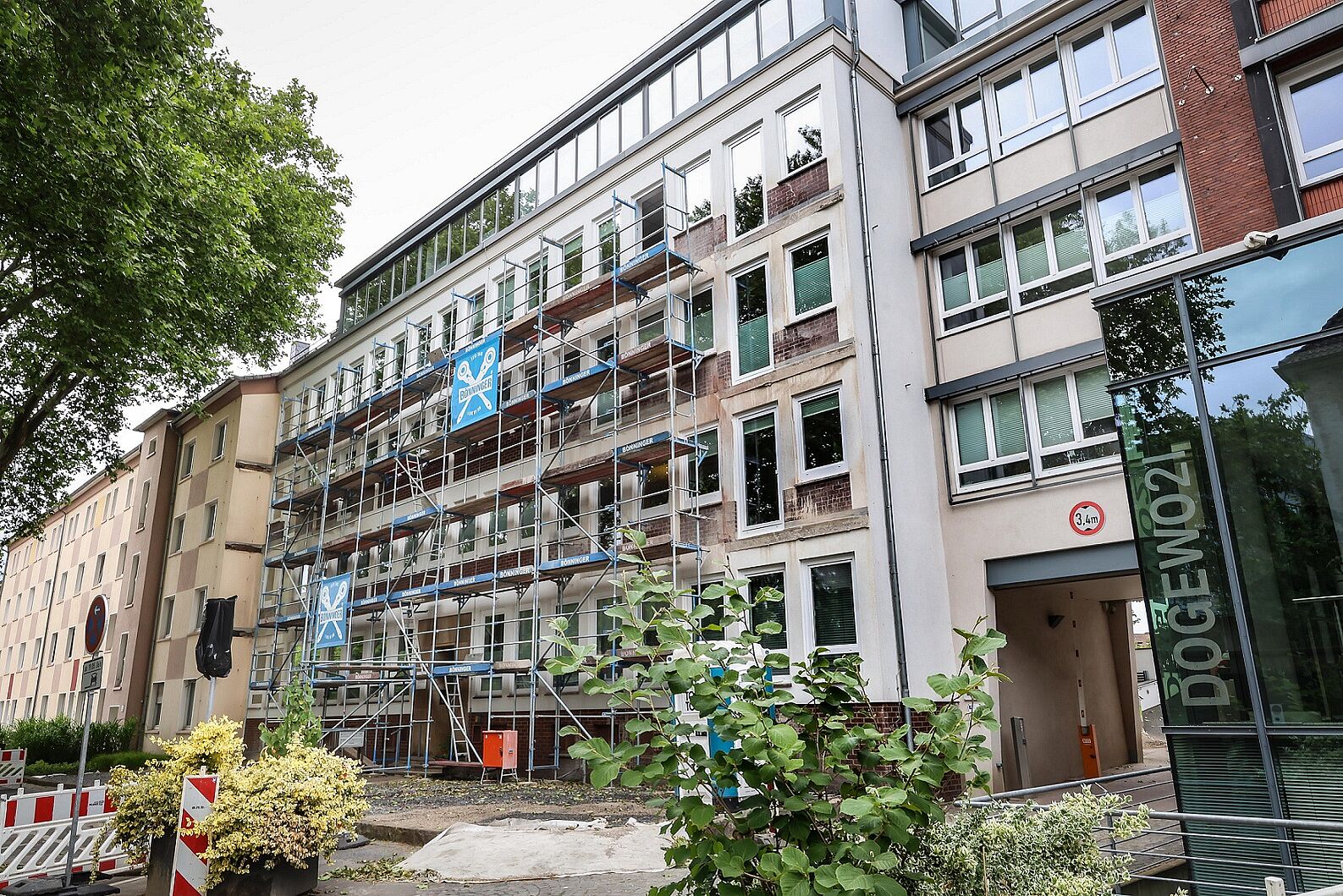 Ein füngeschossiges Verwaltungsgebäude, das teilweise bereits eingerüstet ist und Baustellenbarken auf dem Gehweg..