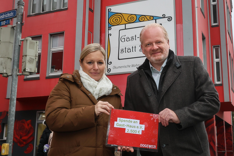 Eine Frau und ein Mann stehen vor einem roten Haus mit einem Schild, auf dem Gast-Haus statt Bank steht, und halten einen symbolischen Spendenscheck in den Händen.