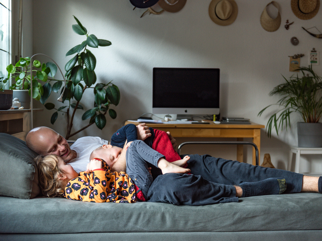 Ein Vater liegt zusammen mit seinen zwei Kindern auf der Couch und kuschelt und lacht mit beiden.