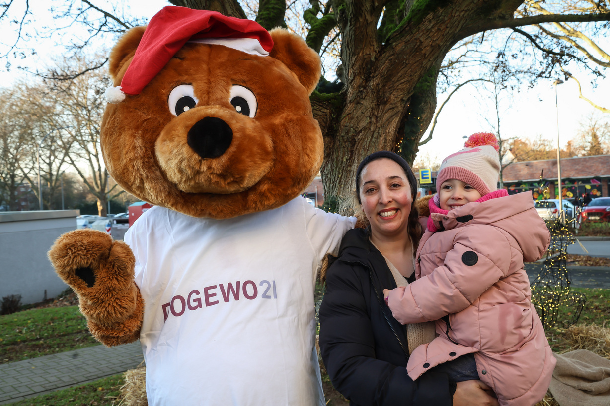 Eine Frau steht mit ihrem Kind auf dem Arm neben dem Maskottchen Dogibär vor einem Baum im Garten des Nachbarschaftstreffs ExtraPlatz.