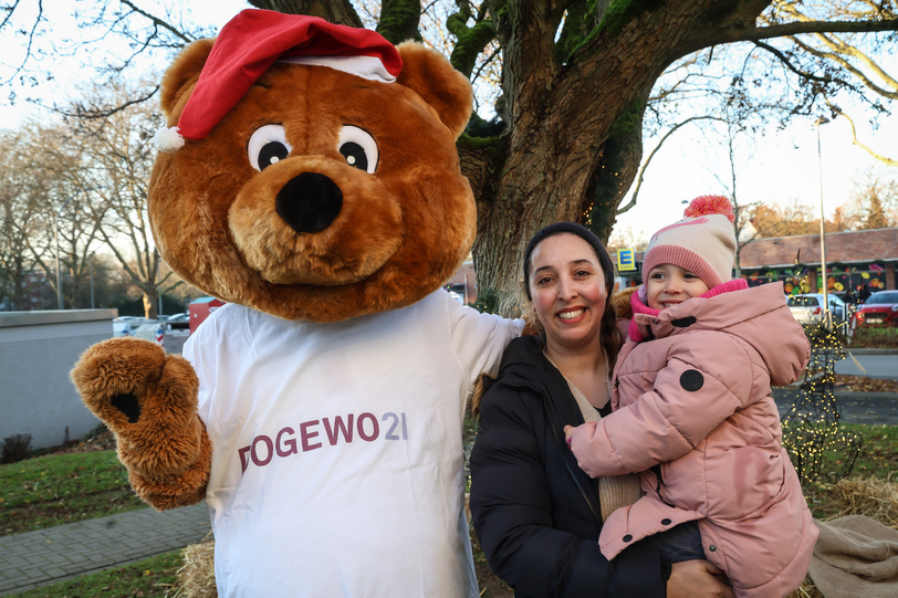 Eine Frau steht mit ihrem Kind auf dem Arm neben dem Maskottchen Dogibär vor einem Baum im Garten des Nachbarschaftstreffs ExtraPlatz.