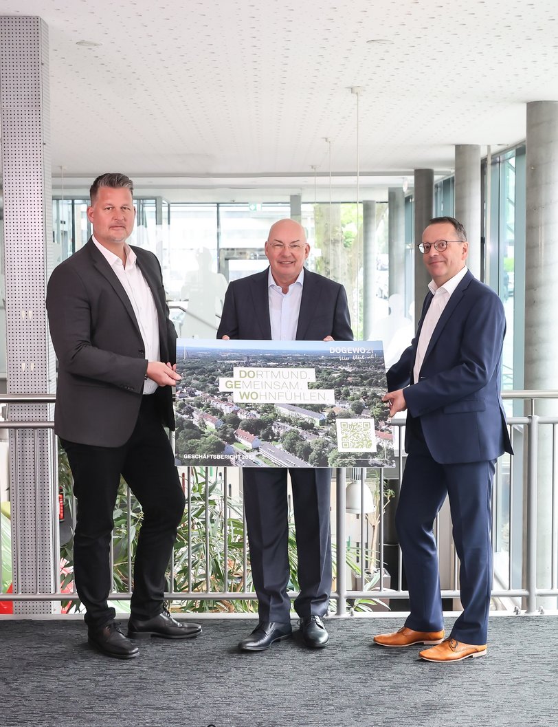 Die drei Herren der Geschäftsleitung von DOGEWO21 präsentieren ein Plakat mit QR-Code und dem Bild des digitalen Geschäftsberichts 2023.