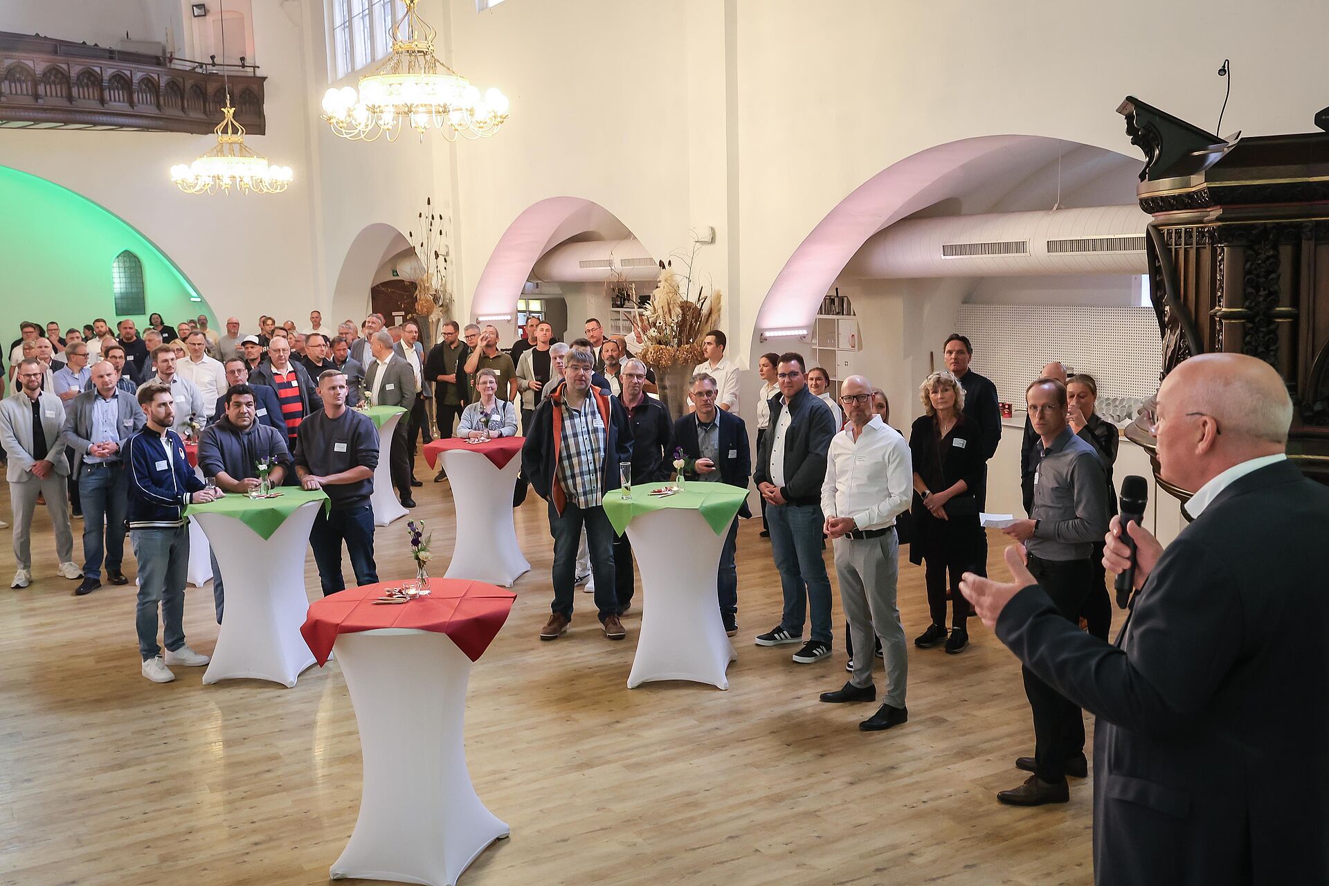 Männer und Frauen stehen in einer Eventkirche bei einer Veranstaltung und hören einem Redner zu.