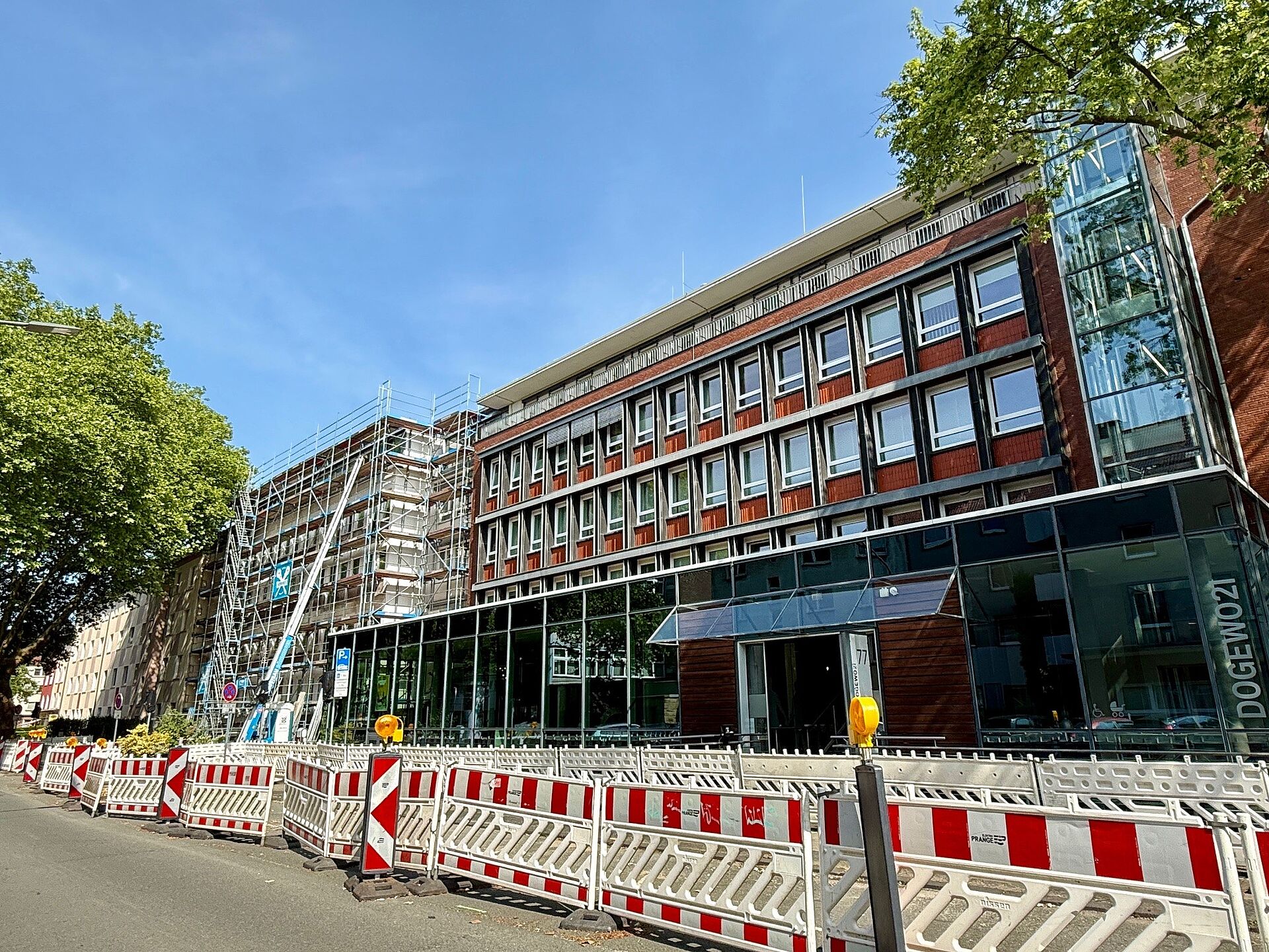 Zwei füngeschossige Verwaltungsgebäude und das gläserne Kundenzentrum, welche teilweise bereits eingerüstet sind, und Baustellenbarken auf der gesperrten Parkfläche davor.