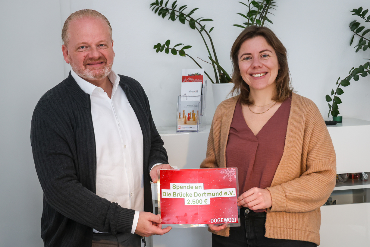 Zwei Menschen stehen mit einem symbolischen Spendenscheck in einem Büro und lächeln in die Kamera des Fotografen.