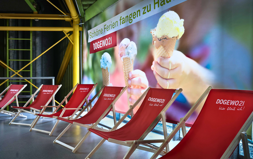DOGEWO21 Liegestühle mit einem Banner im Hintergrund, auf dem Hände mit einer Eiswaffel und tropfendem Eis zu sehen sind.