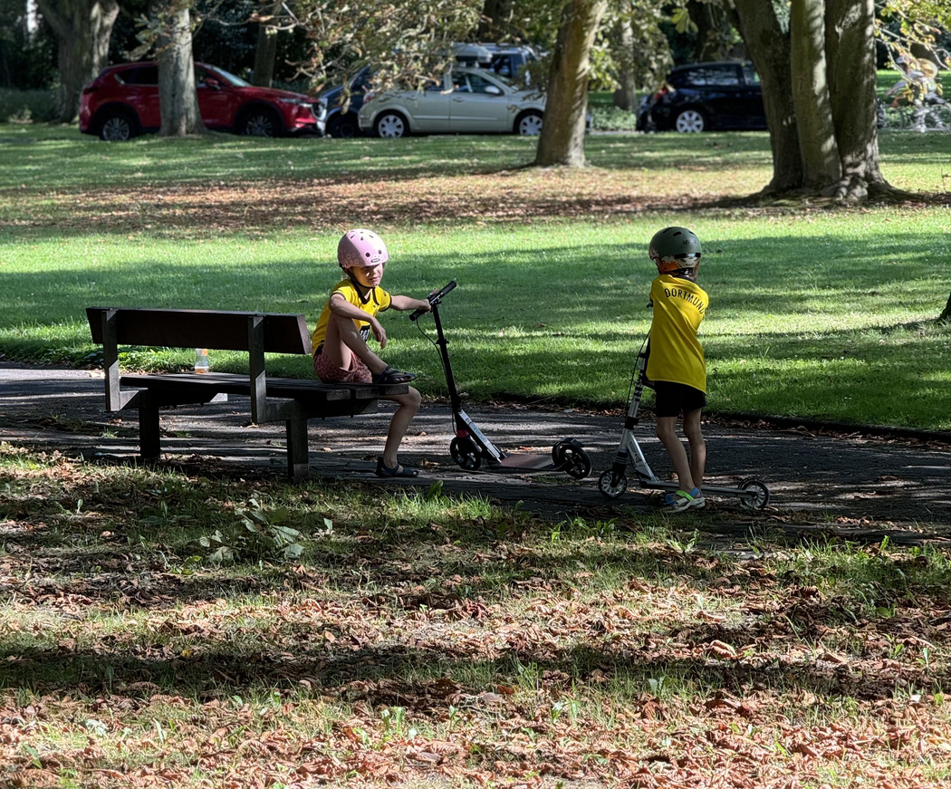 Zwei Kinder mit einem Roller auf einer Bank in einem Park