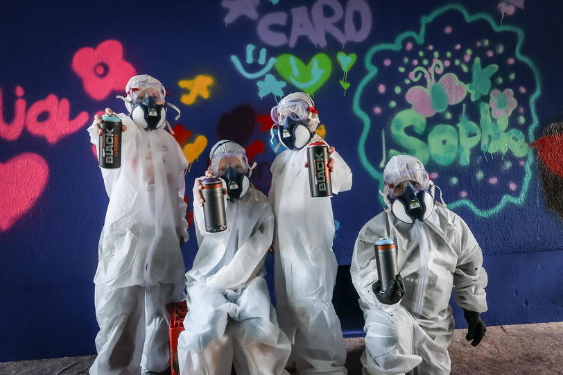 Vier Kinder in Maleranzügen und mit Schutzmasken stehen vor einer Graffitiwand und halten Sprühdosen in die Kamera.
