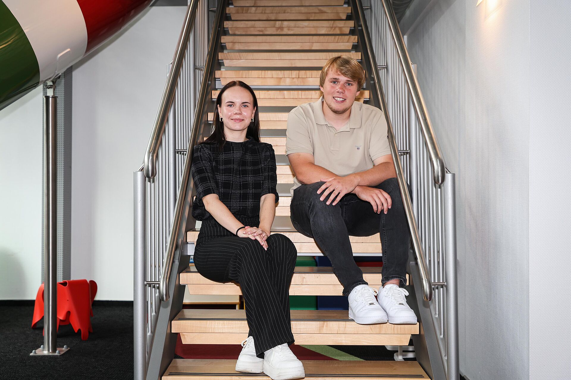 Die beiden neuen Auszubildenden sitzen auf einer Treppe im Kundenzentrum von DOGEWO21 und lächeln in die Kamera