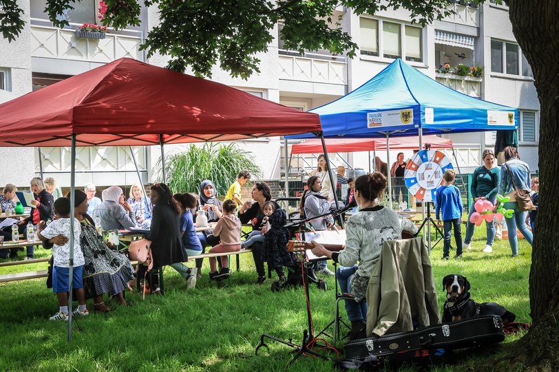 Menschen jeglichen Alters sitzen unter einem Pavillon und singen, essen und reden miteinander während eine Frau mit Gitaree im Vordergrund Lieder spielt.