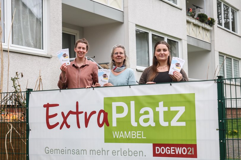 Drei Frauen stehen im Garten des Nachbarschaftstreffs von DOGEWO21, hinter einem Namensbanner, und halten Flyer für eine Seniorensprechstunde in Richtung Kamera.