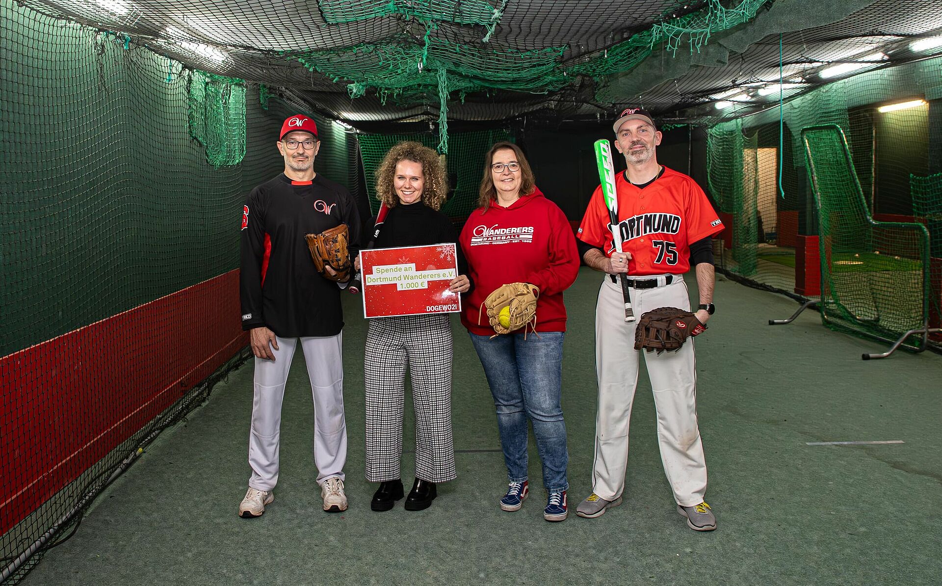 Zwei Frauen und zwei Männer stehen in einer Baseball-Trainingshalle mit Fangnetzen und halten einen symbolischen Spendenscheck.