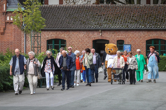 Männer und Frauen gehen über den Weg zum Gelände Zeche Zollern, mittendrin das DOGEWO21-Maskottchen, der Dogibär.