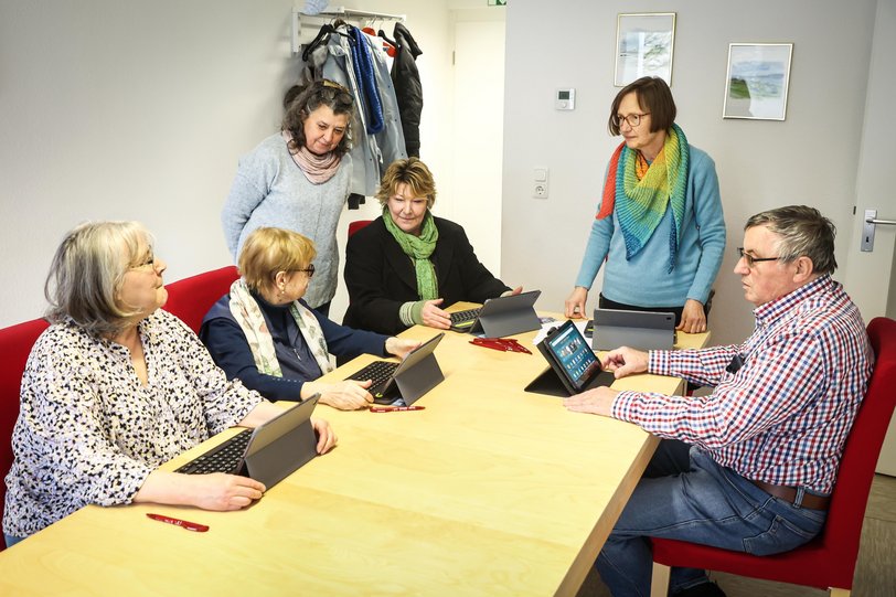 Mehrere Menschen sitzen und stehen interessiert an einem Tisch mit digitalen Tablets