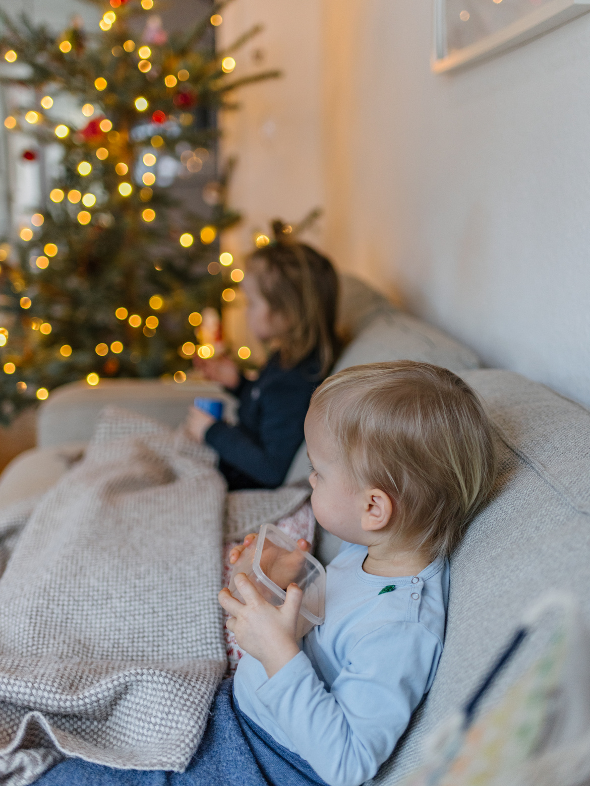 Zwei Kinder mit einer Wolldecke auf der Couch und einer Tasse in der Hand.