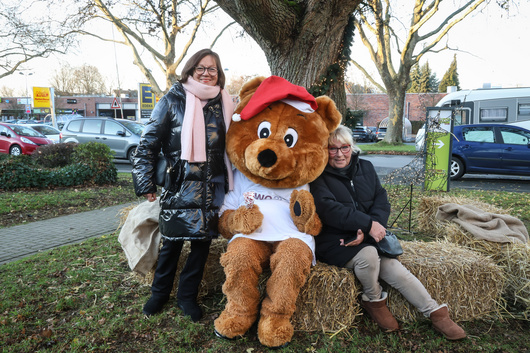 Zwei Frauen lächeln in die Kamera und flankieren das Maskottchen Dogibär im Garten vom ExtraPlatz Wambel.