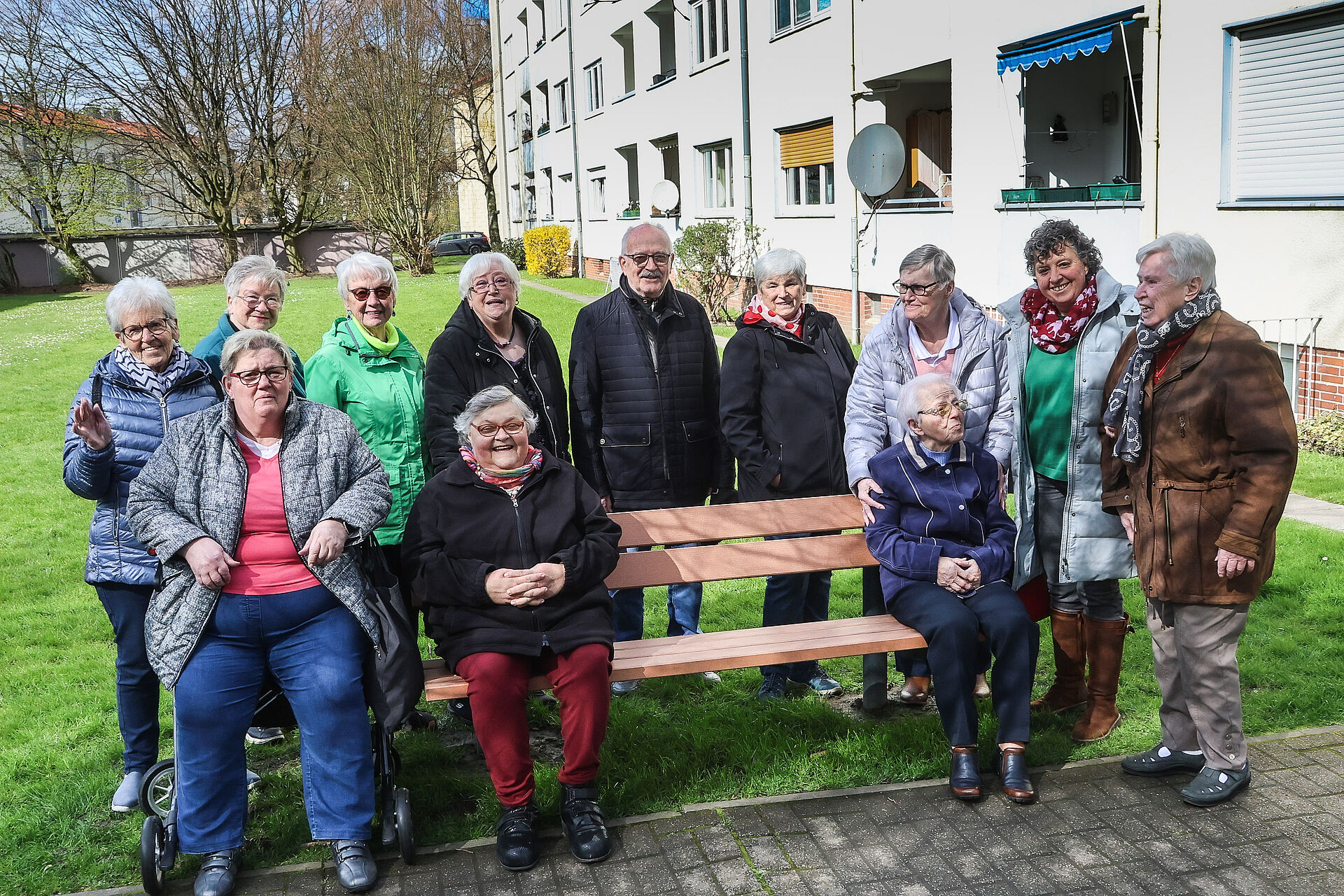 Auf der Grünfläche neben einem Mehrfamilienhaus steht eine Sitzbank aus Holz, auf der mehrere Menschen sitzen und einige Menschen stehen um die Bank herum und schauen in die Kamera 