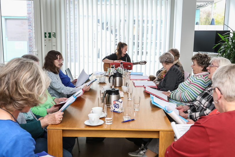 Mehrere Menschen sitzen singend um einen Tisch herum, eine Musikerin begleitet auf der Gitarre,  der Tisch ist gedeckt.