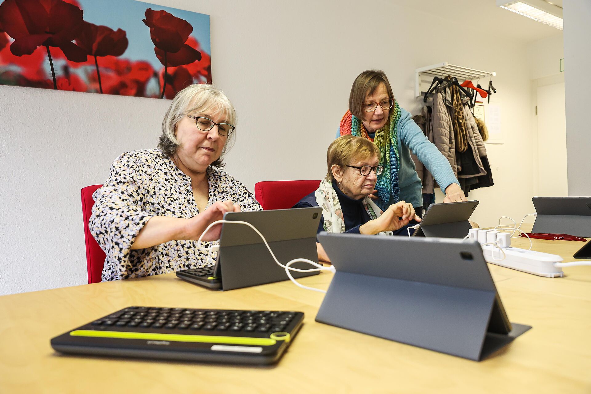 Zwei Frauen sitzen an einem Tisch vor ihren Tablet-PCs und eine dritte Frau erklärt etwas zum Umgang mit dem technischen Gerät