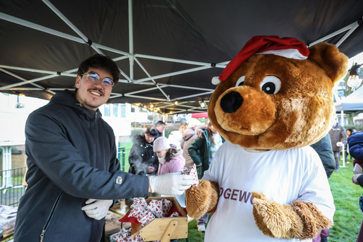 Ein Mann und das Maskottchen Dogibär stehen unter einem Zeltpavillon, in der Hand Tüten mit gebrannten Mandeln.