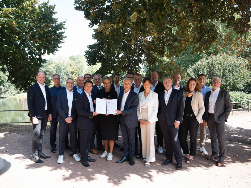 Neunzehn Menschen stehen unter einem Baum, die vorderen Vertreter und die Wirtschaftsministerin halten eine unterzeichnete Absichtserklärung in Richtung des Fotografen. 