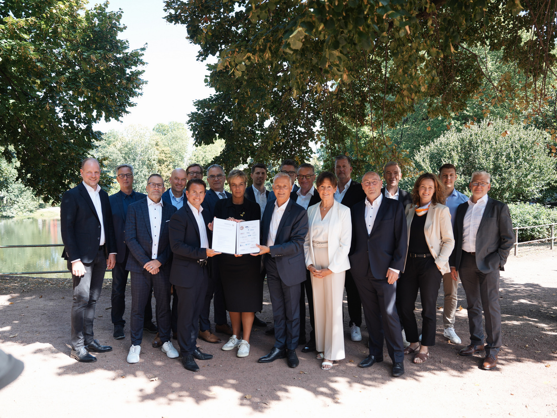 Neunzehn Menschen stehen unter einem Baum, die vorderen Vertreter und die Wirtschaftsministerin halten eine unterzeichnete Absichtserklärung in Richtung des Fotografen.