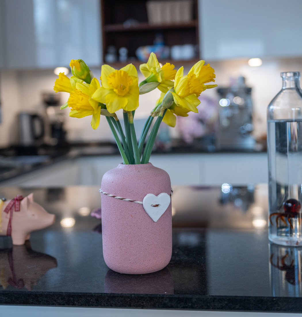 Eine Blumenvase mit Osterglocken und einem Herzanhänger auf einer Küchenablage.