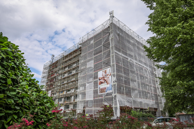 Ein achtstöckiges Mehrparteienhaus ist eingerüstet und mit Schutznetz und DOGEWO21-Bannerversehen, seitlich Büsche und ein Baum.
