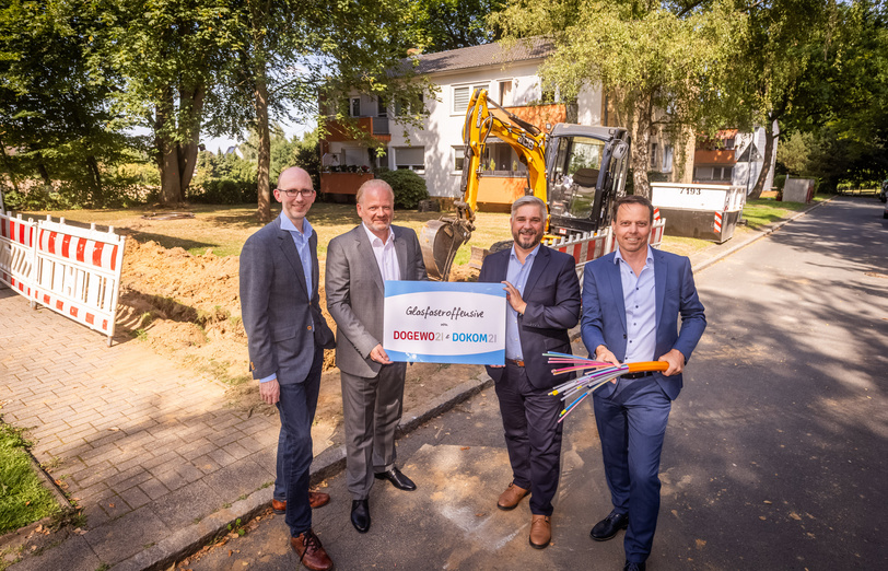 Vier Männer stehen mit einem Schild und Glasfaserkabel neben einer Baustelle zur Verlegung von Kabeln, im Hintergrund ein kleiner Bagger.