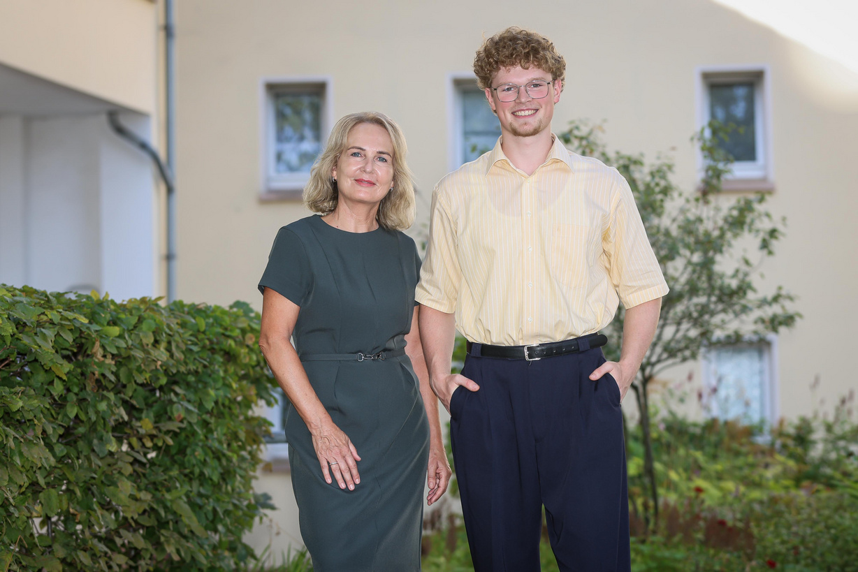 Eine Frau und ein Mann stehen in einem Hof vort einer grünen Hecke und schauen in die Kamera des Fotografen.