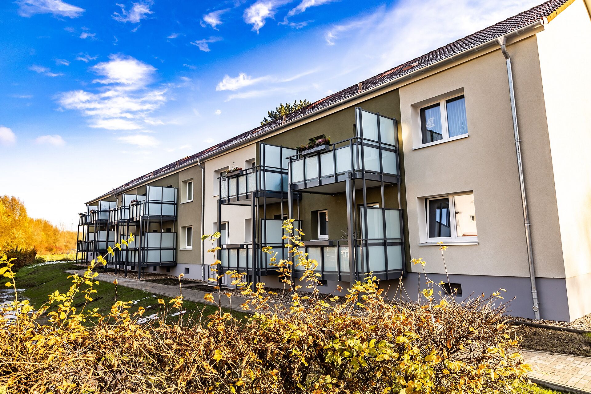 Drei modernisierte Mehrfamilienhäuser mit verputzter Wärmedämmung und mit großen Vorstellbalkonen ist umgeben von Bü+schen und Bäumen unter einem strahlend blauen Himmel