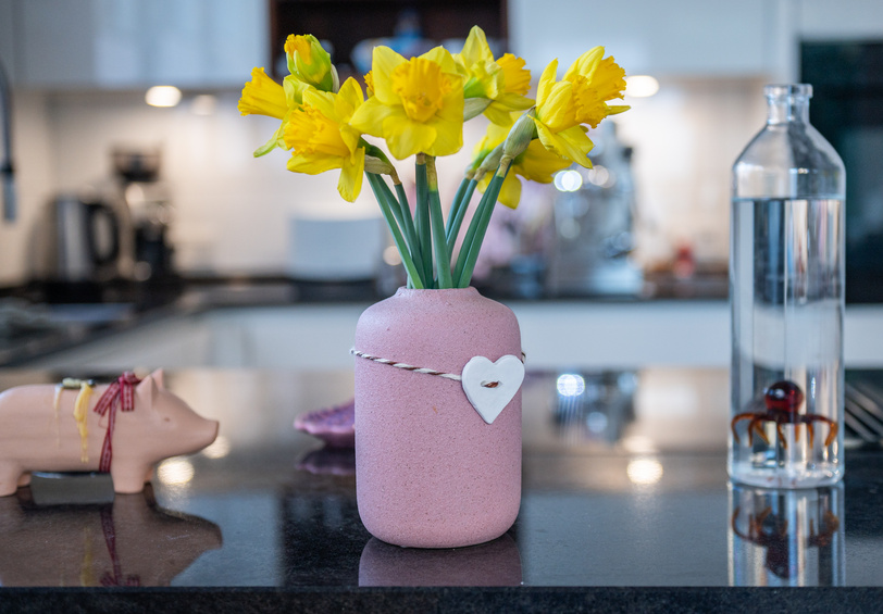 Eine Blumenvase mit Osterglocken und einem Herzanhänger auf einer Küchenablage.