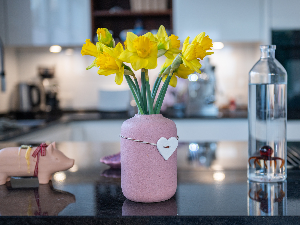 Eine Vase mit Osterglocken und einem Herzanhänger an der Vase. Die Vase steht auf einer Küchenablage.