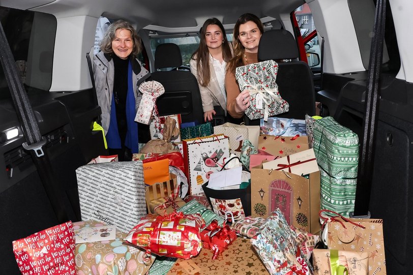 Drei Frauen präsentieren viele bunt verpackte Geschenke, die in einem großen Autokofferraum liegen.