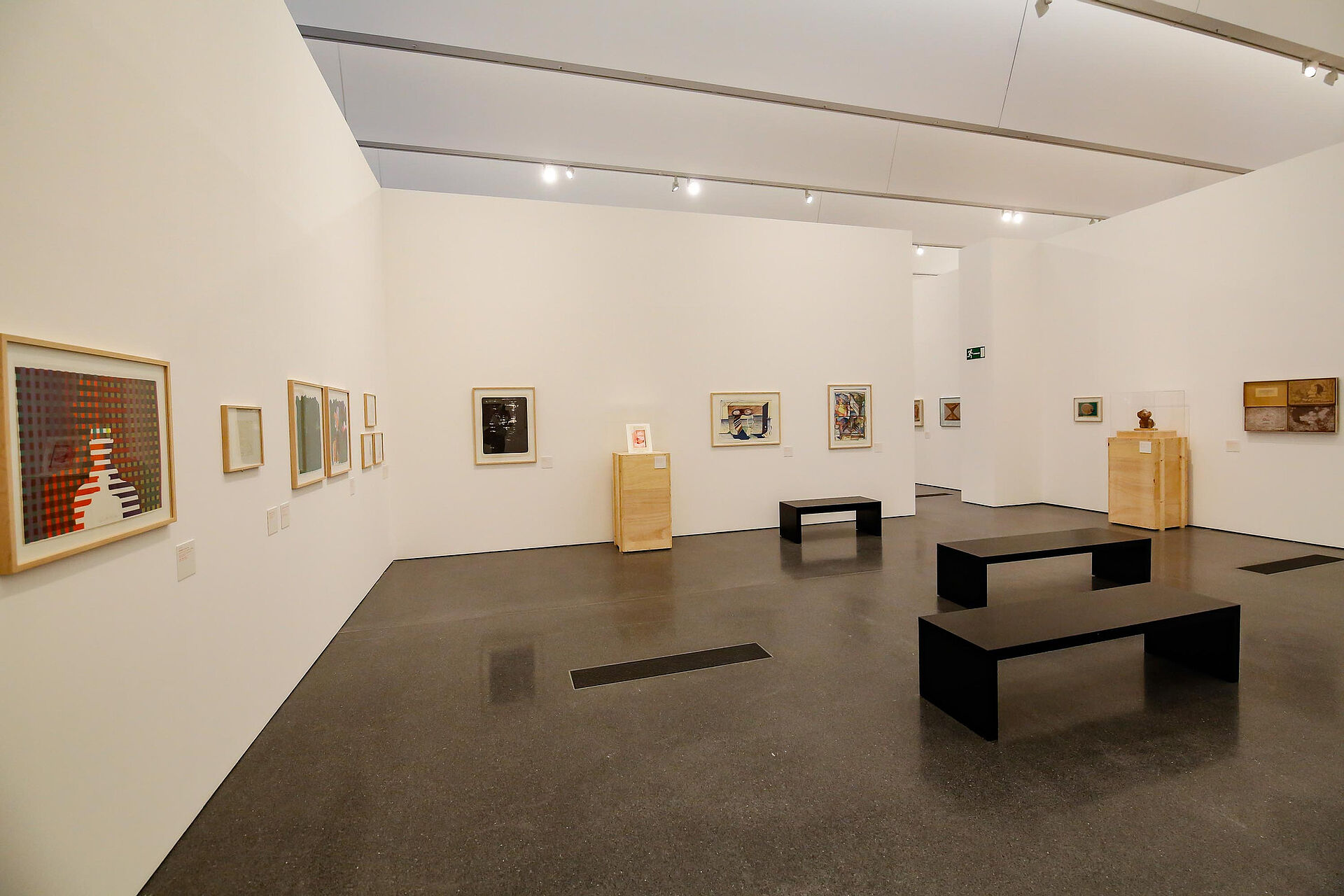 Ausstellungsraum mit wenigen Gemälden, weißen Wänden und drei schlichten schwarzen Bänken in der Mitte des Raumes